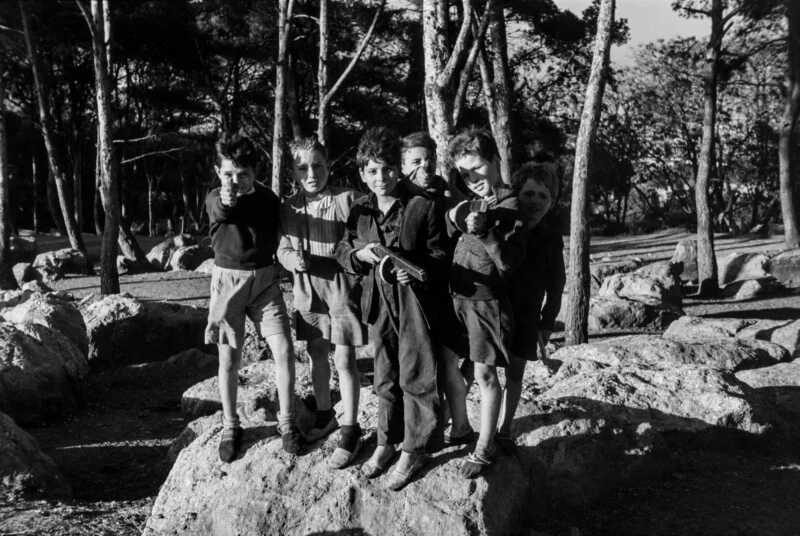 Six young boys stand on a large rock in a wooded area, posing with toy guns pointed toward the camera.