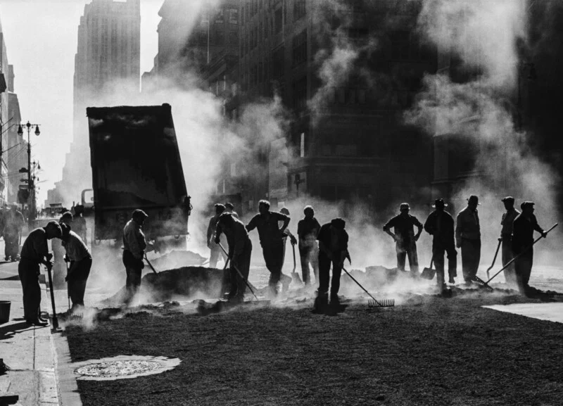 A group of men in work clothes use rakes and shovels to spread asphalt on a city street, surrounded by thick clouds of steam.