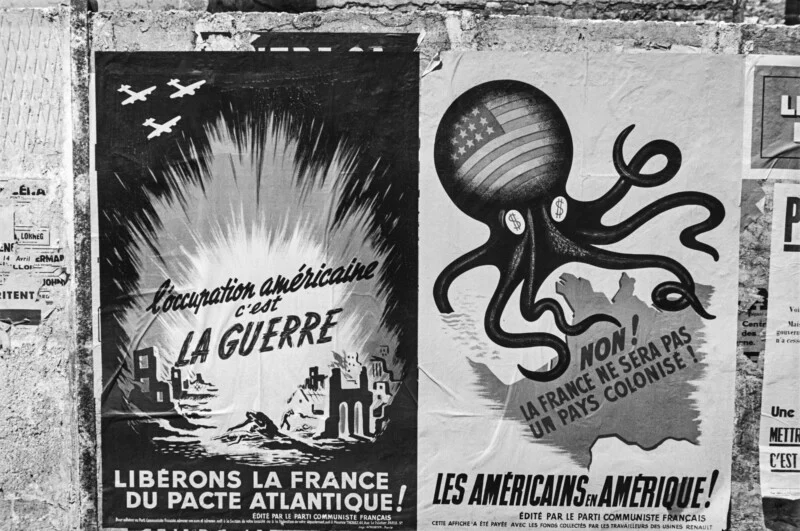 Two French Communist Party propaganda posters on a wall, one depicting war scenes and the other an octopus with a US flag head.