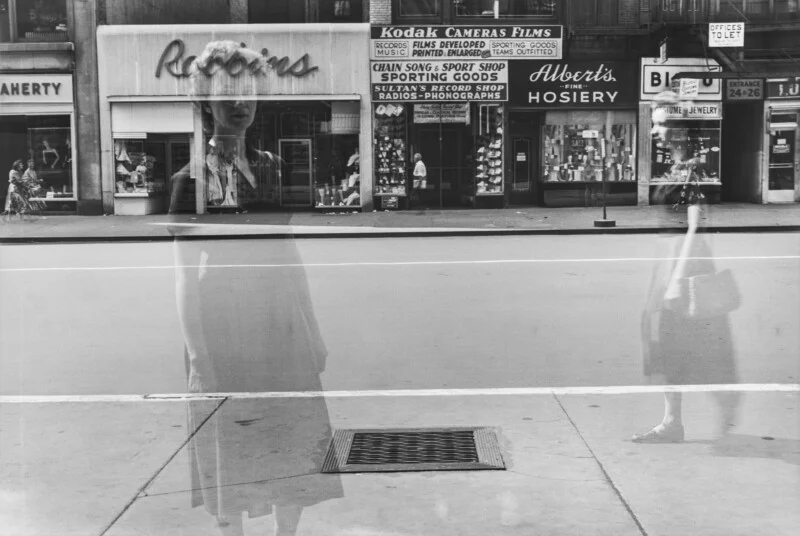 A double-exposure black-and-white photograph shows a street scene with storefronts, including Robbins and Albert's Hosiery, overlaid with ghostly, translucent figures of people walking.