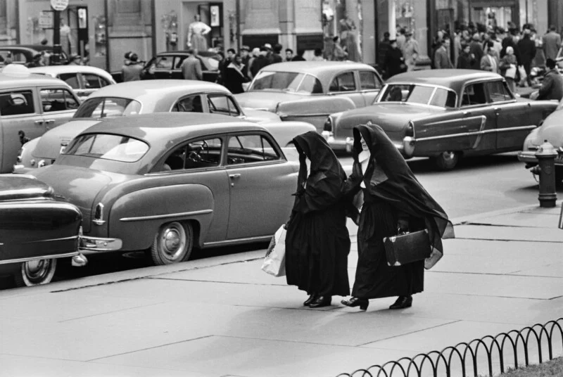 Two nuns in black habits walk along a city sidewalk past a row of parked vintage cars.
