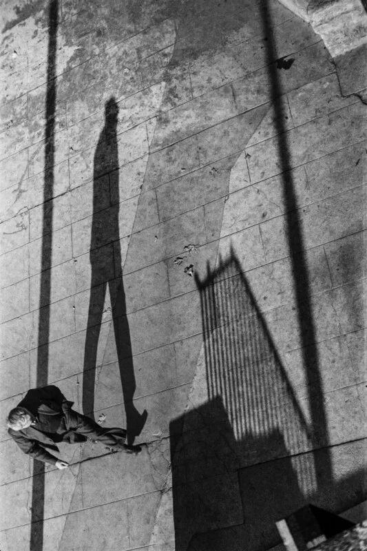 A high-angle, black-and-white shot shows a man walking on a paved surface, casting a long, elongated shadow beside a fence.