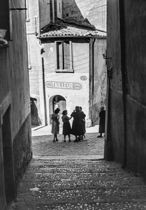 A group of women stands in a cobblestone square framed by narrow alley walls, with a building in the background labeled