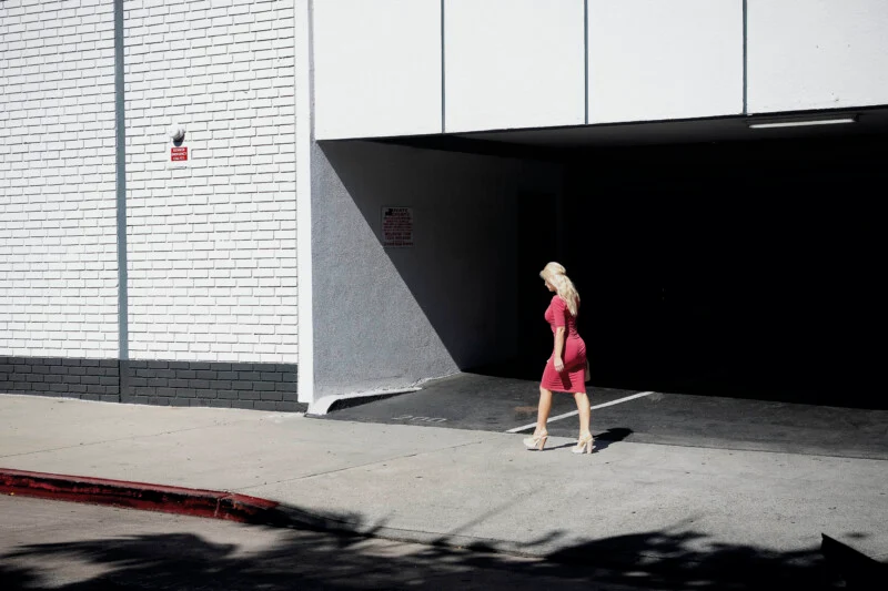 A blonde woman in a pink dress and high heels walks past the entrance of a parking garage on a sunny day.