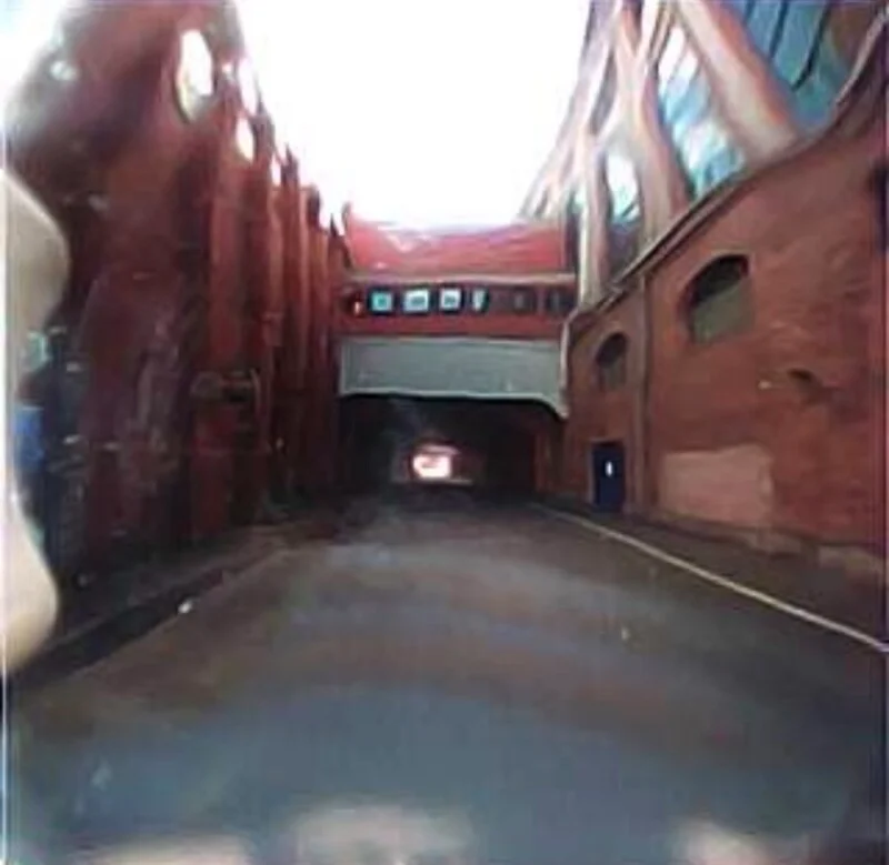 A narrow road passes beneath a red brick building connected by an elevated walkway between two industrial structures.