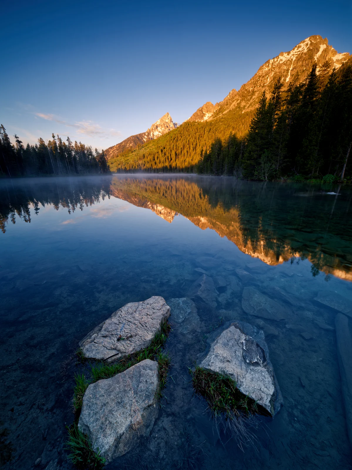 Sunlight illuminates a mountain peak and forest reflected in a calm, clear lake with large rocks in the foreground.