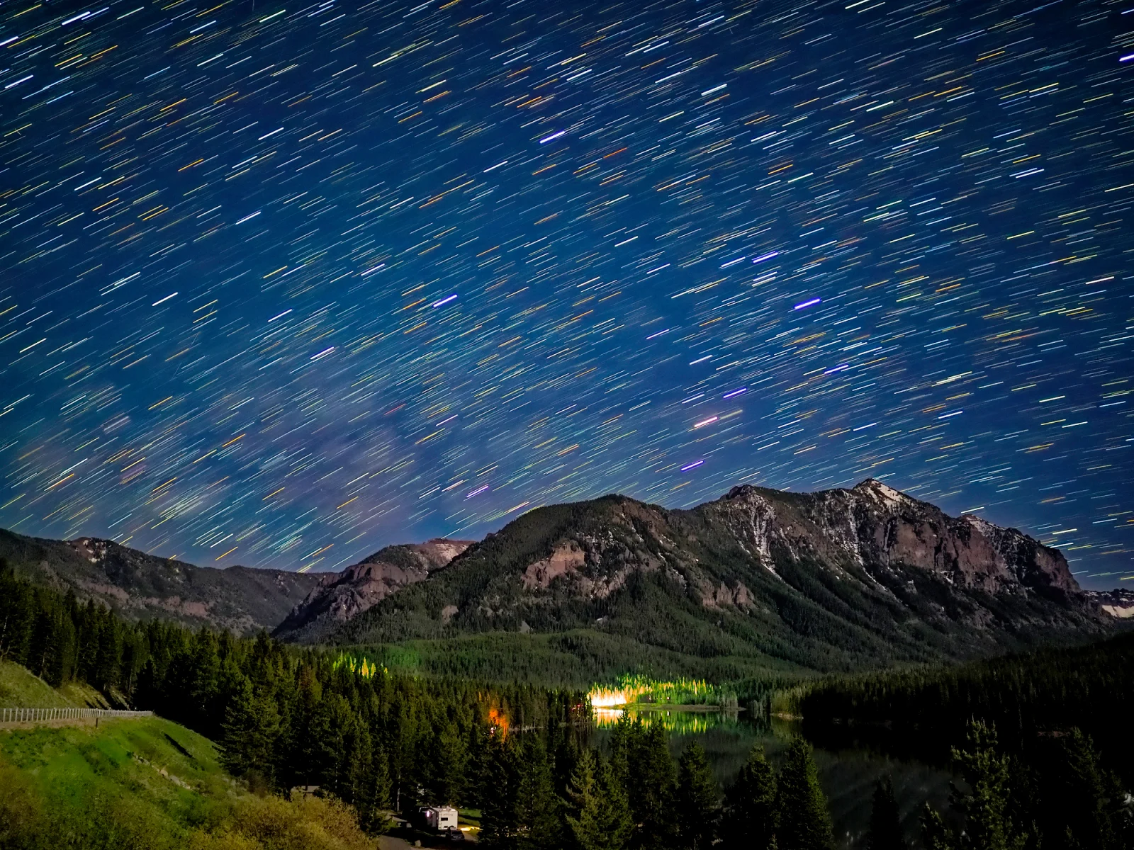 A long-exposure night shot shows star trails streaking across a dark blue sky above a mountain range and a forested lake.