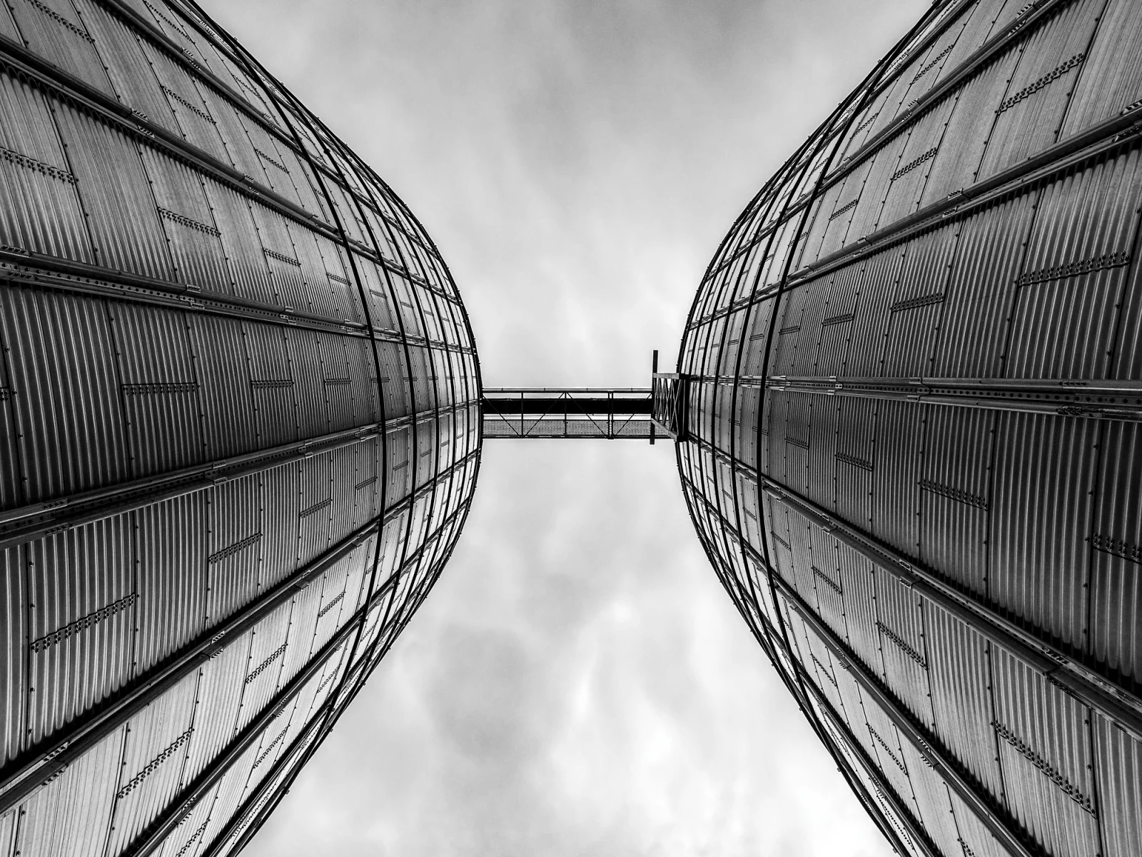 A low-angle, black-and-white view of two large, corrugated metal grain silos connected by a narrow bridge against a cloudy sky.