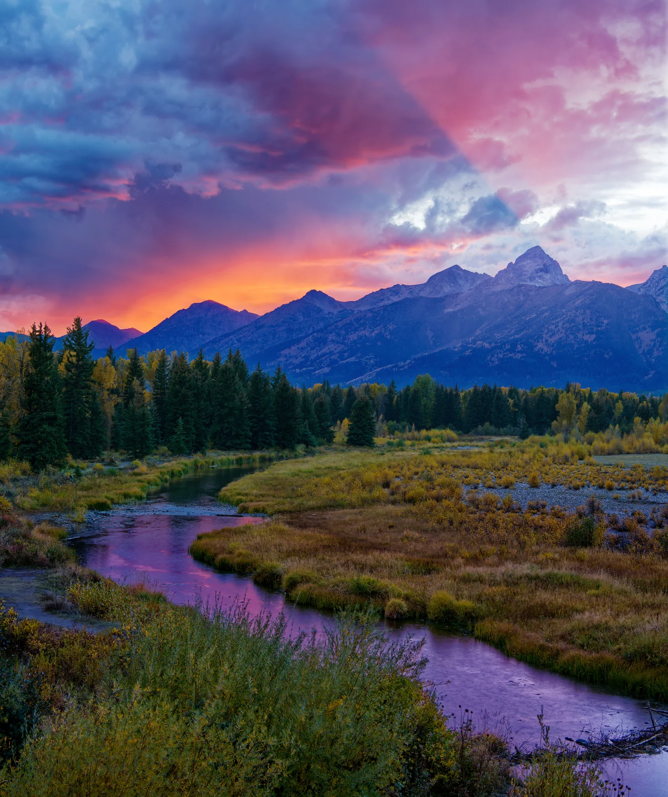A winding river flows through a grassy meadow toward a forest and a jagged mountain range under a vibrant sunset sky.