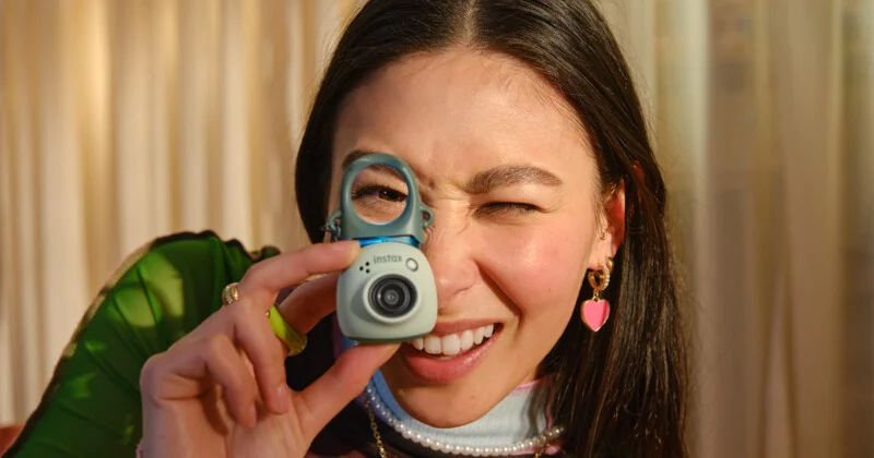 A person with long dark hair winks while holding a small, light green digital camera up to one eye.