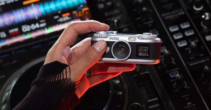 Een hand met een zwarte mouw houdt een zilveren en zwarte Instax-camera vast voor een DJ-mixingconsole.