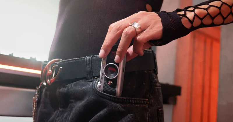 Een persoon met een zwarte top en spijkerbroek met een visnetmouw houdt een kleine zilveren camera gedeeltelijk in de zak.