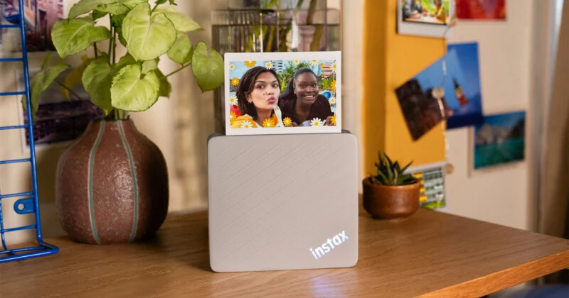 A light gray Instax printer sits on a wooden desk with a small photo of two women emerging from the top, next to a potted plant and a blue wire rack.