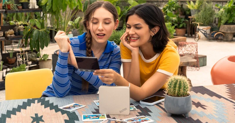 Two young women sit at a table outdoors, smiling as they look at a small, rectangular photo print held between them.