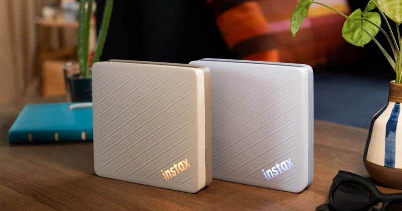 Two portable, square-shaped electronic devices with diagonal textured surfaces and illuminated "instax" logos sit on a wooden table next to a potted plant and sunglasses.