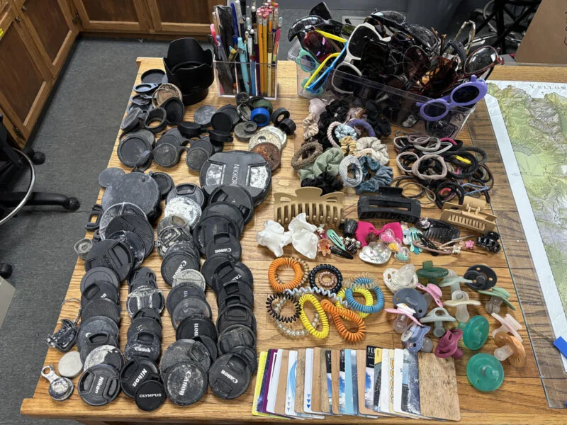 A wooden table covered in a collection of lost and found items, including lens caps, sunglasses, hair accessories, pacifiers, and a row of plastic cards.