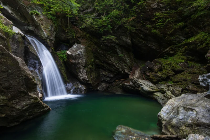 Een waterval die in een diepe, groene poel stort, omgeven door mosachtige rotsformaties en dicht bos.