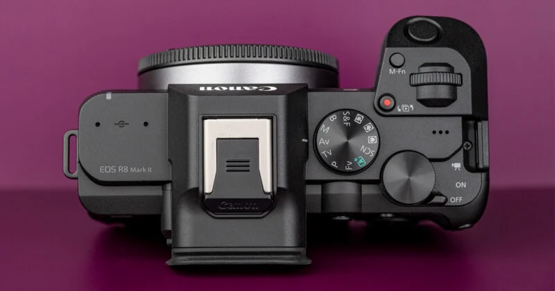 Bovenaanzicht van een zwarte, spiegelloze camerahoes met een moduledraaiwiel, sluiterknop en hotshoe tegen een paarse achtergrond.