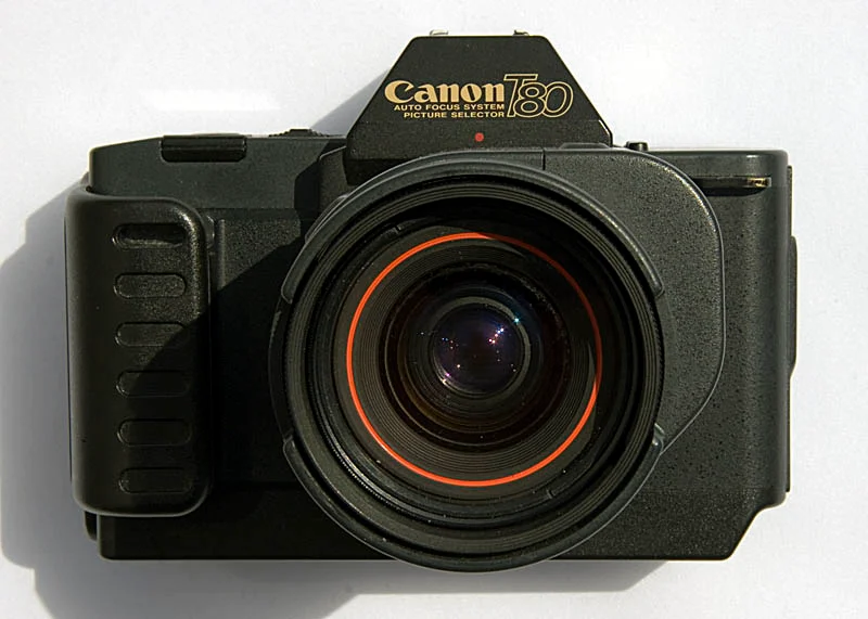 Een zwarte Canon T80 35mm filmcamera met een erop gemonteerde lens, gezien vanaf de voorkant tegen een witte achtergrond.