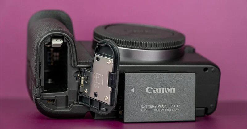 Een Canon LP-E17-accupakket ligt naast de geopende batterijdeur van een zwarte digitale camera tegen een paarse achtergrond.
