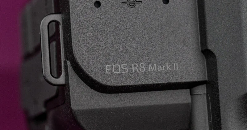 Een zoom-in op een zwarte camerabehuizing met de tekst 'EOS R8 Mark II' op de zijkant, naast een metalen polsriemhaak.