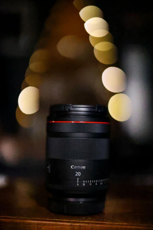 Een zwarte Canon-lens met een rode ring staat rechtop op een houten ondergrond, met een achtergrond van vervagende, gouden bokeh-lichten.