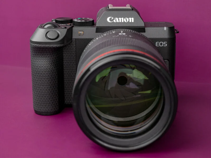 Een zwarte Canon EOS-mirrorless camera met een grote lens gemonteerd, tegen een purperen achtergrond.