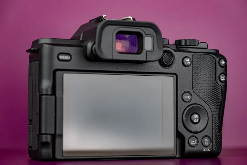 Achterkant van een zwarte digitale spiegellose camera met een groot LCD-scherm, zoeker en diverse knoppen tegen een felle magenta achtergrond.