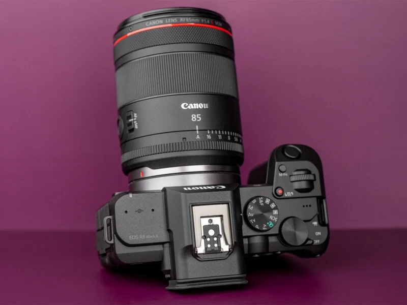 Een Canon EOS R8 Mark II-camera met een Canon RF 85mm-lens bevestigd, tegen een paarse achtergrond.
