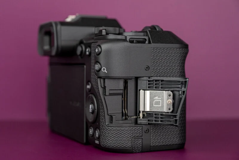 Een zwarte digitale camera wordt vanaf de achter- rechterkant getoond, met een geopende batterijdeur, waardoor het interne compartiment zichtbaar is.