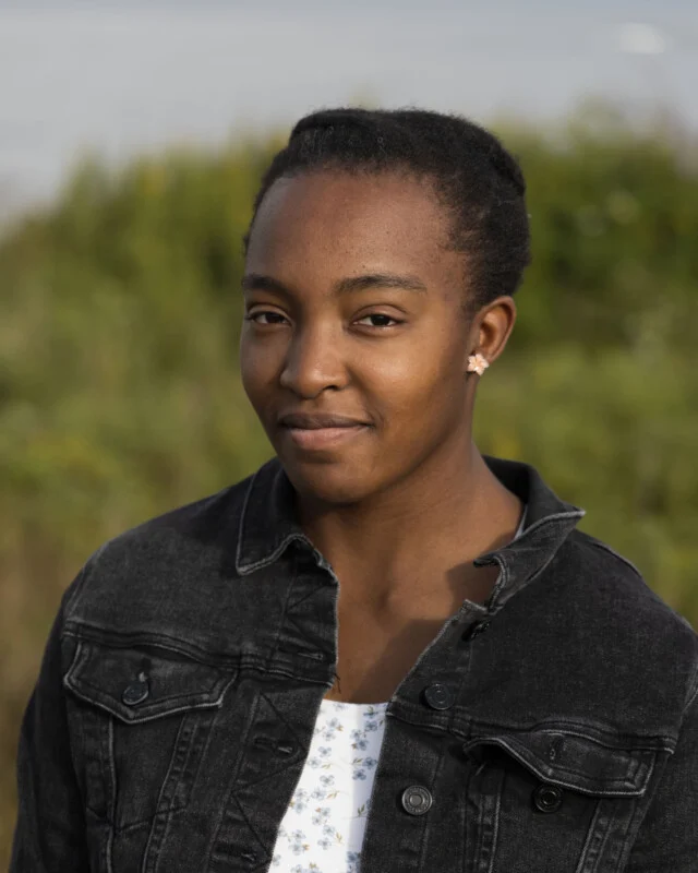 Een jonge vrouw met kort donker haar en een lichte bloemige top onder een zwarte spijkerjas kijkt naar de camera tegen een vervaagde groene achtergrond.