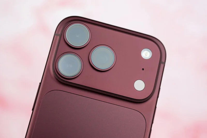 Een close-up van de achterkant van een donkerrode smartphone, met een drievoudige lens-camera, een flitser en een microfoon ingebouwd in een verhoogd rechthoekig module.