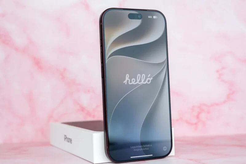 Een donkergekleurde smartphone staat rechtop op zijn witte winkeldoos tegen een roze marmeren achtergrond, met een welkomstscherm in het Hongaars weergegeven.