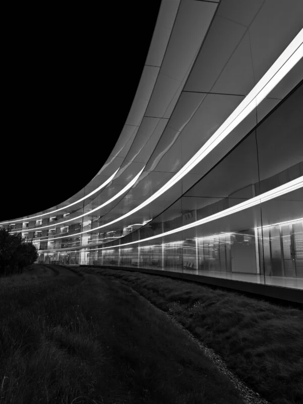 Een zwart-wit lage hoekopname van een gebogen, moderne glazen gebouw bij nacht, met horizontale lichtstroken en een groenachtig landschap op de voorgrond.