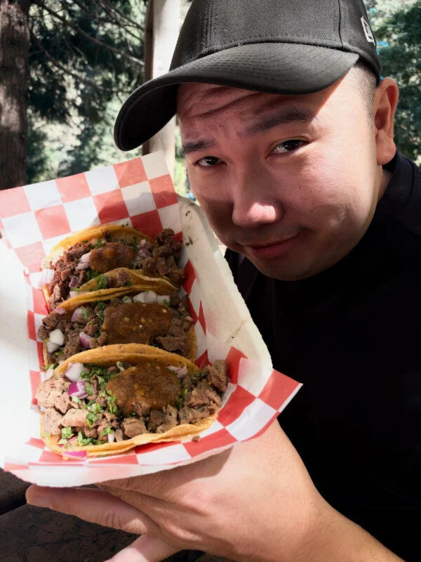 Een man met een zwarte pet houdt een rood-wit geruite papieren bordje met drie taco’s bovenop, belegd met vlees, ui, koriander en salsa.