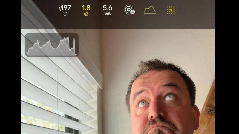 Een man kijkt omhoog in een kamer met een raam, terwijl een camera-interface met een histogram-overlay zichtbaar is bovenaan.