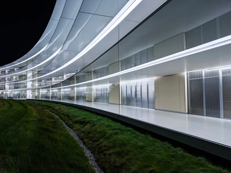 Een gewelfde, moderne gebouw met glazen wanden en verlichte horizontale lichtstroken kijkt uit over een gazon met een helling.' width=