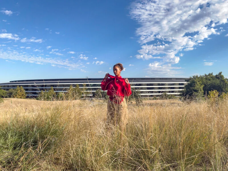 Een persoon met krullend haar draagt een rode blouse en bruine broek in een veld van hoog, droog gras voor een lang modern gebouw onder een blauwe lucht met verspreide wolken.