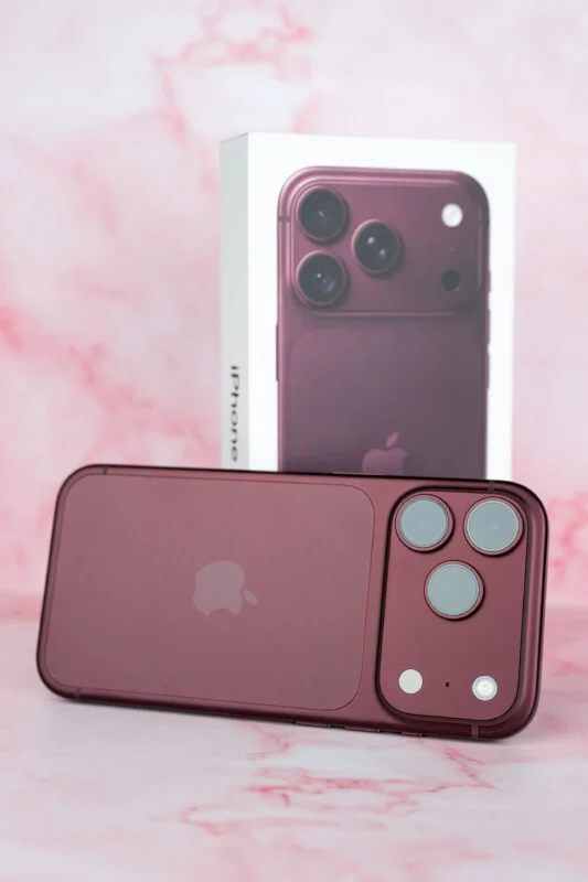 Een donkerrode smartphone ligt horizontaal voor zijn bijbehorende productdoos op een roze marmeren oppervlak.