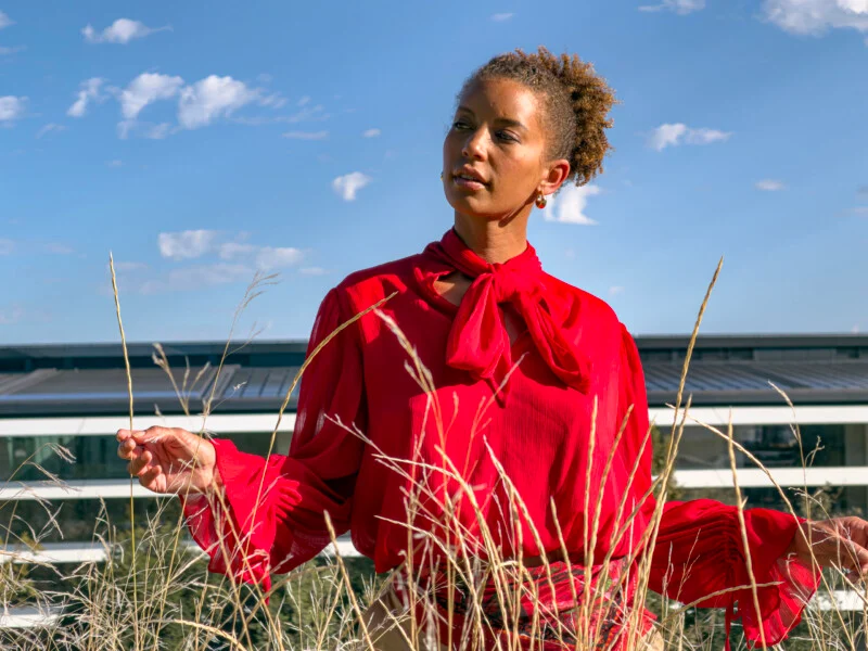 Een vrouw met krullend haar draagt een rode blouse met een strik in de hals terwijl ze in een veld met hoog gras staat tegen een blauwe lucht en een moderne gebouw.