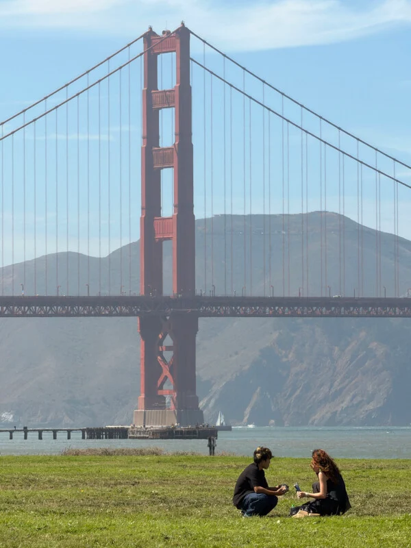 Twee personen zitten op een gazon voor de Golden Gate Bridge onder een heldere blauwe lucht.