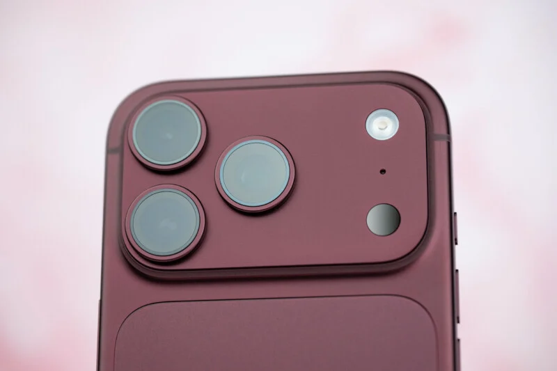 Een close-up van de achterkant van een donkerrode smartphone, met een drievoudige lens-camera, een flitser en een klein sensor op een verhoogd rechthoekig module.
