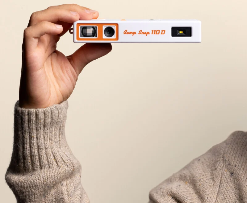 A hand in a beige sweater holds a white and orange Camp Snap 110 D digital camera against a plain light background.