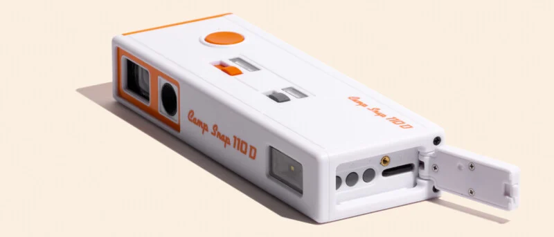 A white and orange Camp Snap 110 D digital camera sits on a light surface with its side port door open, revealing a memory card slot.