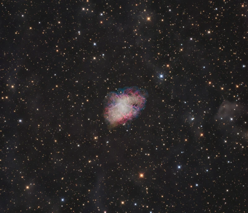 The Crab Nebula, a colorful, filamentary supernova remnant, sits centered against a dense, dark field of distant stars.