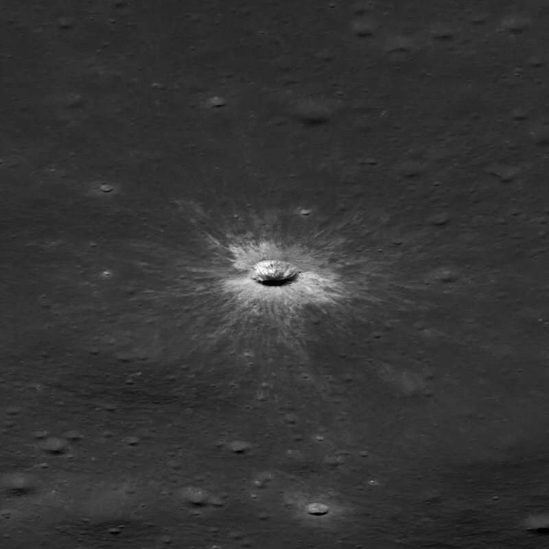 A small, bright impact crater with a prominent radial ray system sits in the center of a dark, smooth lunar surface.