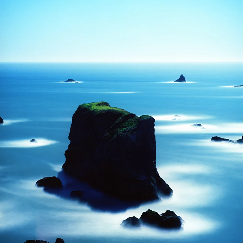 A large, moss-covered rock formation stands in the center of a calm, blue ocean with smaller rocks scattered around it.