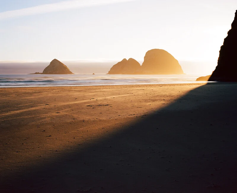 A sandy beach at sunset with large rock formations in the distance and a long shadow cast across the foreground.