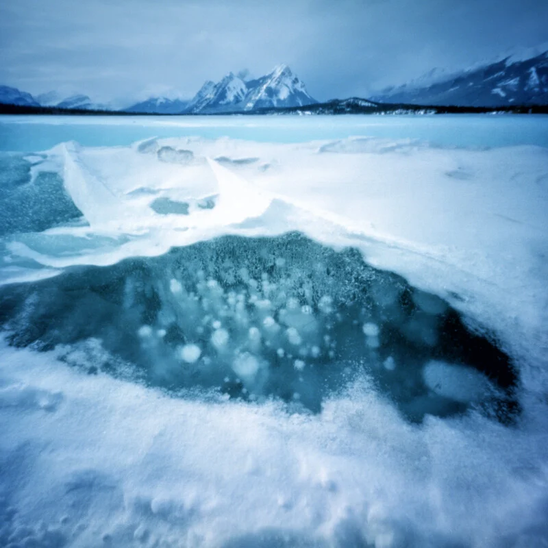 A patch of clear ice on a frozen lake reveals trapped air bubbles, with snow-covered ice and distant mountains in the background.