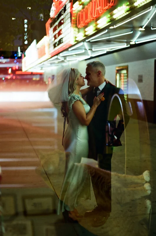 A bride and groom stand close together, facing each other in front of a brightly lit theater marquee at night.